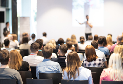 Back of an audience looking towards a speaker giving a talk at an event.