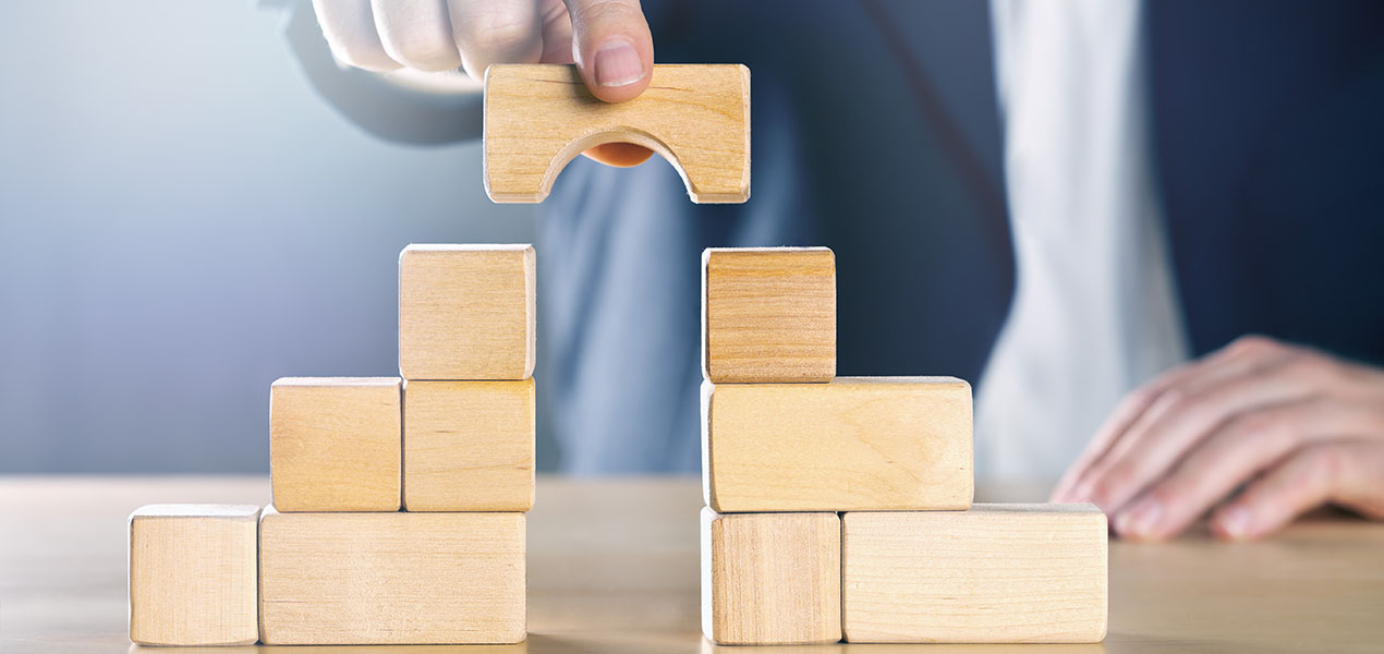 Person in suit adding a wood block to bridge the gap between two towers of wooden blocks.