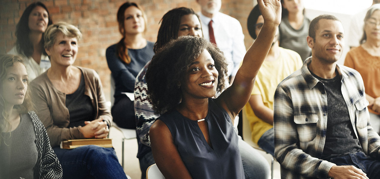 An audience of business people looking towards a speaker, with one girl in the audience raising her hand.